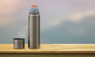 Termos, kahveli metal bardak ve ahşap masa, bulanık doğa arka planı, yakın plan.