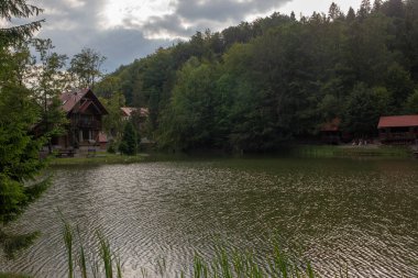 Bereketli yeşil ormanların ve bulutlu bir gökyüzünün altında büyüleyici ahşap kulübelerin arasına kurulmuş sakin bir göl barışı çağrıştırıyor..
