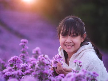 Beyaz önlüklü, pırıl pırıl gülümseyen küçük bir kız margaret çiçekleri tarlasında duruyor.