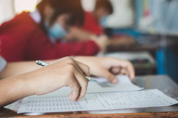 University student holding pen or pencil writing on paper answer sheet sitting on chair lecture final exam participates in exam room or classroom in uniform student