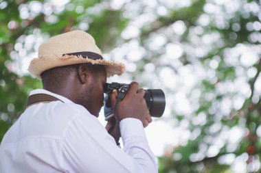 Şapkalı ve beyaz gömlekli Afrikalı bir adam tatil sırasında elinde bir kamera, seyahat ve fotoğraf konseptiyle gezintiye çıkıyor.