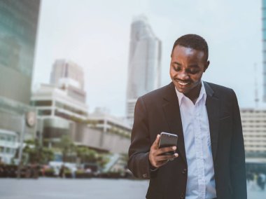 Başarılı Afrikalı iş adamı, modern ofis binasının dışında telefonu kullanıyor takım elbiseli akıllı adam gülümsüyor ve akıllı telefon ekranına bakıyor.