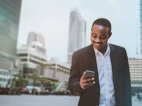 Successful african businessman using phone outside modern office building, Smart man in business suit smiling and looking at smartphone screen texting