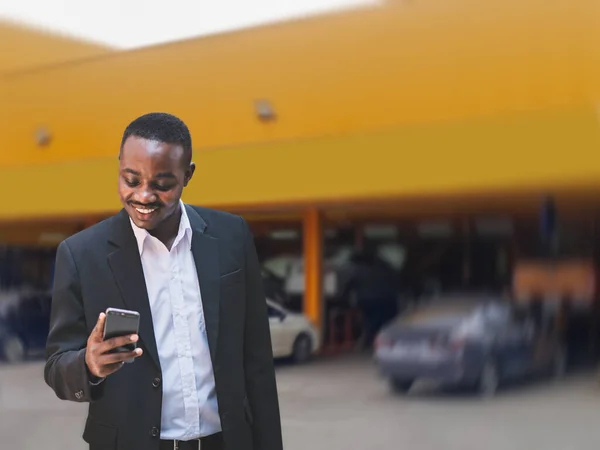 African businessman takes his car for repairs at a modern auto repair shop. A customer comfortably looks at his smartphone while waiting for his car to be repaired
