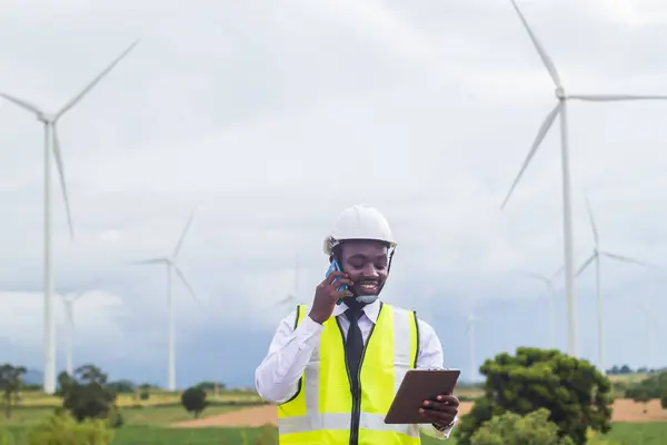 African engineer man stands with smile front the wind turbines generating electricity power station.Concept of sustainability development by alternative energy