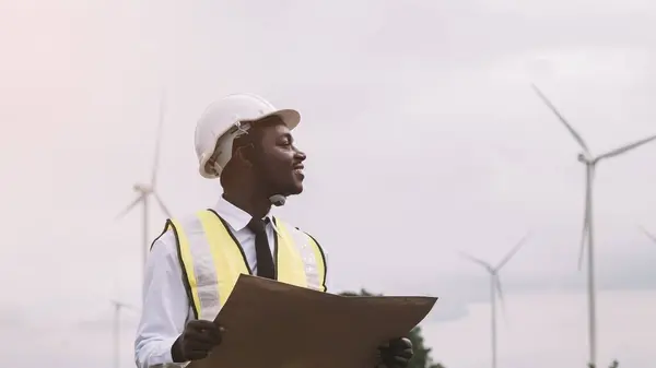 Successful african engineer man stands with smile front the wind turbines generating electricity power station.Concept of sustainability development by alternative energy
