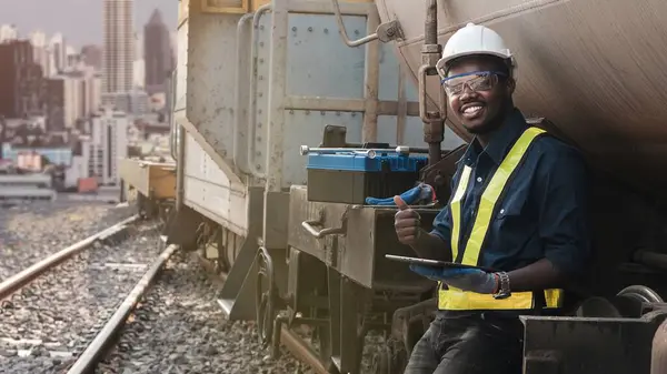 African mechanical engineer looks at tablet to maintain train in city center with tall buildings in the background
