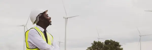 African engineer stand with smile near the wind turbine for inspect the operation of large wind turbines that converts wind energy into electrical energy