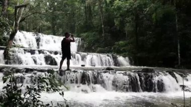 Sırt çantalı gezgin, inanılmaz tropikal şelalenin keyfini çıkarıyor. El kaldırıyor, seyahat ediyor ve tatil kavramını başarıyor.