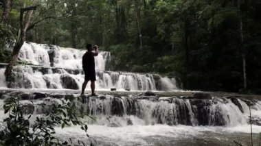 Sırt çantalı gezgin, inanılmaz tropikal şelalenin keyfini çıkarıyor. El kaldırıyor, seyahat ediyor ve tatil kavramını başarıyor.