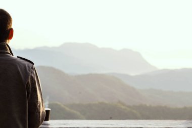 Palto giymiş bir gezgin, ahşap bir balkonda bir fincan sıcak kahveyle güzel bir dağ manzarasına bakıyor..