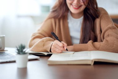 Close up - Hand of woman writing in spiral notepad placed on wooden desktop with various items at home. copy space.