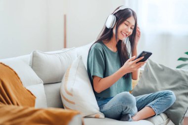 Mutlu Asyalı kadın evdeki kanepede oturmuş cep telefonundan müzik dinliyor, sabah kulaklıkla gülümseyen kız, dinlenme zamanı. Boşluğu kopyala.