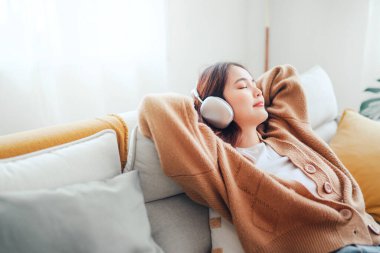 Mutlu Asyalı kadın evdeki kanepede oturmuş cep telefonundan müzik dinliyor, sabah kulaklıkla gülümseyen kız, dinlenme zamanı. Boşluğu kopyala.