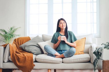 Happy young asian woman drinking coffee relaxing on sofa at home. Smiling female enjoying resting sitting on couch in modern living room.