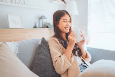 Neşeli genç Asyalı kadın evdeki kanepede otururken bir fincan kahvenin keyfini çıkarıyor, rahat bir sahne, kışın sıcak çay içen güzel bir kadın..
