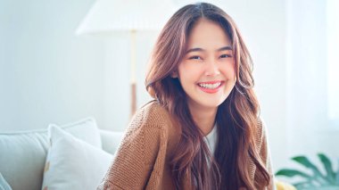 Confident smiling young adult asian woman looking at camera while sitting at home, Happy beautiful lady pretty face dental smile posing alone indoors.
