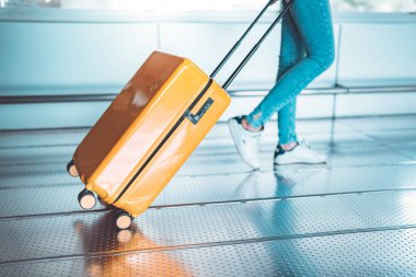 Genç bir bayan yolcu, modern havaalanı terminalinde sarı bir bavulla yürüyor, yolcu kapısına doğru giden kadının arka görüntüsü, seyahat ya da tatil yolculuğuna hazır.