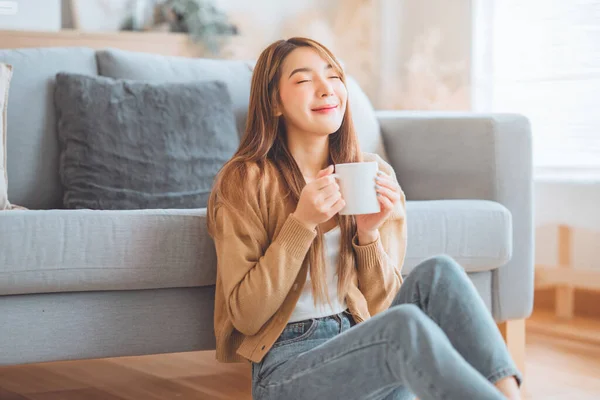 stock image Joyful young asian female enjoying a cup of coffee while sitting on the rug beside to the sofa at home, Cosy scene, Smiling pretty woman drinking hot tea in winter.