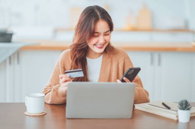 Çevrimiçi ödeme, mutlu Asyalı kadın kredi kartı tutuyor ve online alışveriş için akıllı telefon ve dizüstü bilgisayar kullanıyor.
