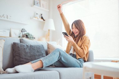 Heyecanlı genç Asyalı kadın telefonda flört uygulamasında sohbet ediyor. Mutlu hissediyor. Kanepede oturuyor, kapalı kapılar ardında sevinçli bir kadın telefonda iyi haberler okuyor. Şaşkın kadın telefonda zaferi kutluyor.