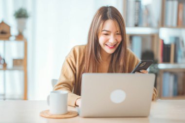 Smiling young asian woman using laptop while holding a cup of coffee and smartphone at home. lifestyle