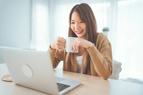 在家里拿着一杯咖啡 一边用笔记本电脑笑着年轻的亚洲女人 生活方式 图库图片
