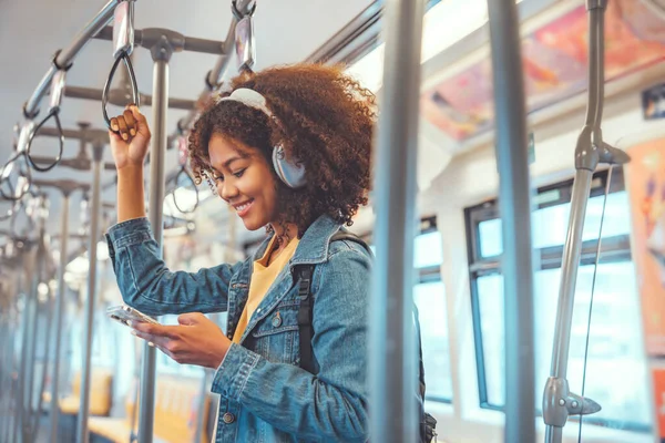 快乐的年轻非洲裔美国女性乘客在火车上通过智能手机听音乐 微笑的女性在乘坐电车 生活方式和交通工具时佩戴无线耳机 图库照片