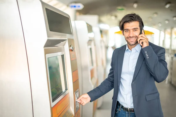 The caucasian man uses a transport card at the ticket machine for travel by sky train in the rush hour time while he uses a smartphone for talking with his friend.