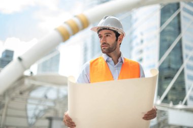 Elinde büyük bir gazete tutan genç beyaz bir adam açık mavi gömlek ve turuncu yelek giyen bir adam inşaat alanında güvenlik için beyaz kask takıyor..