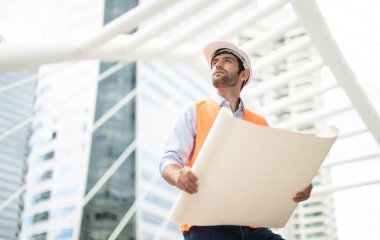 Elinde büyük bir gazete tutan genç beyaz bir adam açık mavi gömlek ve turuncu yelek giyen bir adam inşaat alanında güvenlik için beyaz kask takıyor..