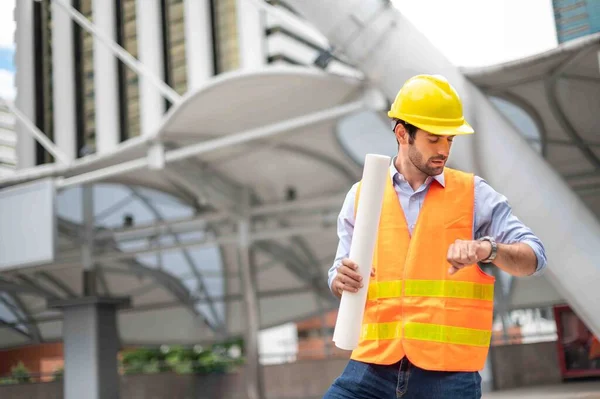 Young caucasian man holding a big paper, guy wearing light blue shirt and jeans with orange vest and yellow helmet for security in construction area, while he looking at his hand watch.