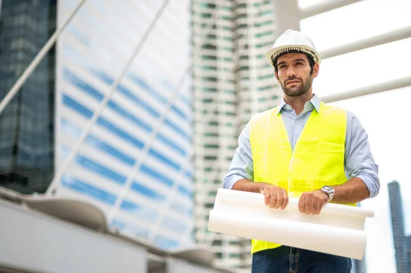 stock image Caucasian man engineer , wearing yellow vest and big hard hat, and hand holding the white floor plan in the site work of the center city.