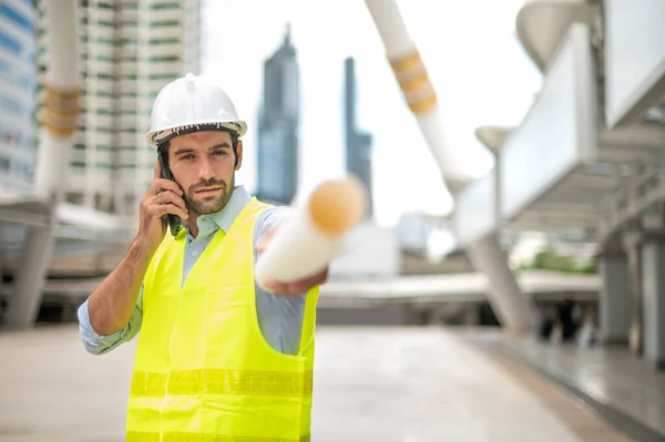 Caucasian man engineer use a smartphone for talking, wearing yellow vest and big hard hat, and the other hand holding the white floor plan in the site work of the center city.