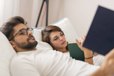 Aşık bir çift, uyandıktan sonra yatakta uzanıyor, sarılıyor ve kitap okuyor.