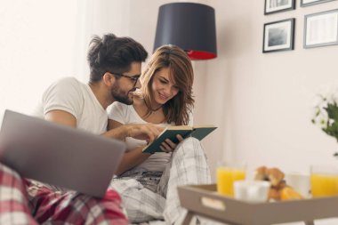 Genç ve güzel bir çift yatakta kahvaltı ederken internette kitap okuyup sörf yaparken eğleniyor. Kıza odaklan.