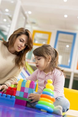 Anne ve kızı bir oyun odasında oturmuş renkli plastik yapı taşlarıyla oynuyorlar. Kızına odaklan.