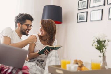 Genç ve güzel bir çift yatakta kahvaltı ederken internette kitap okuyup sörf yaparken eğleniyor. Kıza odaklan.