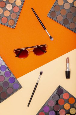 Renkli arka planda çeşitli makyaj ürünlerinin düz yatışı. Makyaj fırçaları, göz falı paletleri, ruj ve güneş gözlüğü