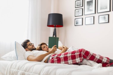 Aşık bir çift, uyandıktan sonra yatakta uzanıyor, sarılıyor ve kitap okuyor.