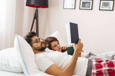 Aşık bir çift, uyandıktan sonra yatakta uzanıyor, sarılıyor ve kitap okuyor.
