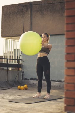 Binanın çatısındaki terasta duran bir kadın, pilates topuyla çalışıyor.