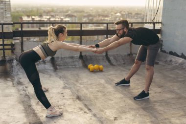 Bir binanın terasında duran genç çift, el ele tutuşup antrenmandan önce esniyorlar; arka planda kentsel ufuk çizgisi
