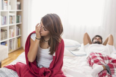 Yatakta tartıştıktan sonra üzgün bir çift; erkek arka planda kızgın yatarken kadının kafası karışık bir şekilde oturuyor.