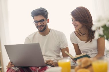 Genç ve güzel bir çift yatakta kahvaltı ederken internette kitap okuyup sörf yaparken eğleniyor. Adama odaklan.