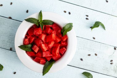 Karpuz tohumları ve nane yapraklarıyla dekore edilmiş açık mavi bir arka planda bir kâse karpuz dilimleri.