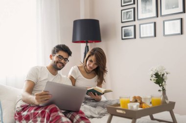 Genç ve güzel bir çift yatakta kahvaltı ederken internette kitap okuyup sörf yaparken eğleniyor. Kıza odaklan.