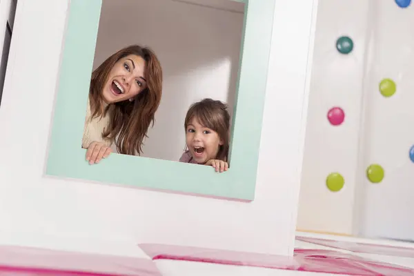 快乐的母亲和女儿躲藏在一个小木屋里的游戏室里 透过窗户偷窥 做鬼脸 — 图库照片