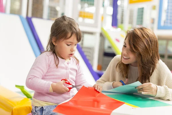 Mother and daughter sitting on a playroom floor, cutting colorful collage paper with scissors and having fun. Focus on the daughter