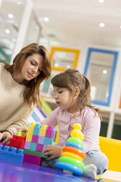 Mother and daughter sitting on a playroom floor, playing with colorful plastic building blocks. Focus on the daughter